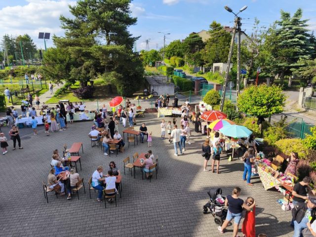 Zdjęcia z lotu ptaka na plac szkolny, na którym odbywa się szkolny festyn. Pośrodku stoliki z krzesełkami, naokoło stoika ze smakołykami. W oddali rozgrywki na boisku szkolnym.