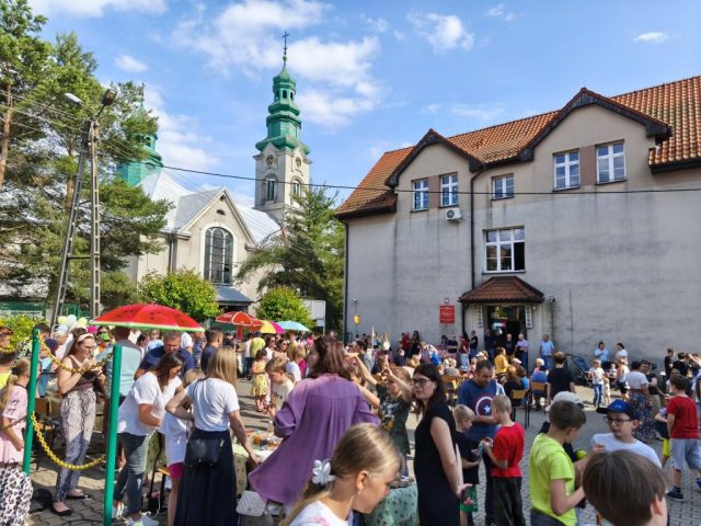 Uczniowie i społeczność lokalna na placu szkolnym podczas rodzinnego festynu. Naokoło placu stoiska ze smakołykami. W tle fasada budynku szkoły, w oddali kościół.