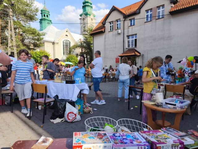Uczniowie i społeczność lokalna na placu szkolnym podczas rodzinnego festynu. Na środku placu stoły z krzesełkami, zaś naokoło placu stoiska ze smakołykami.
