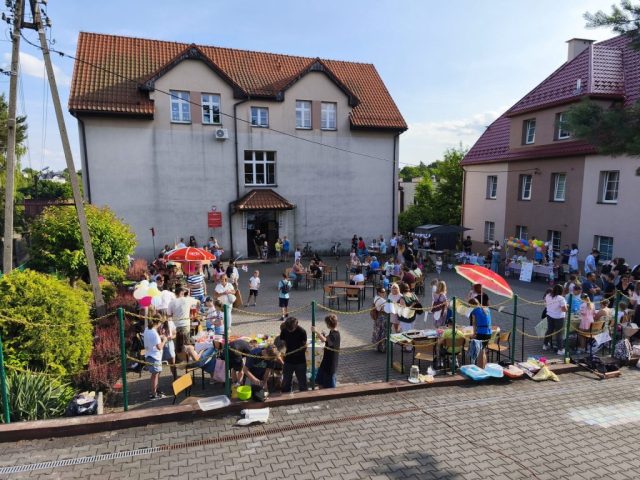 Widok na plac szkolny, na którym rozłożono stoiska z owocowymi smakołykami. W tle budynek szkoły.