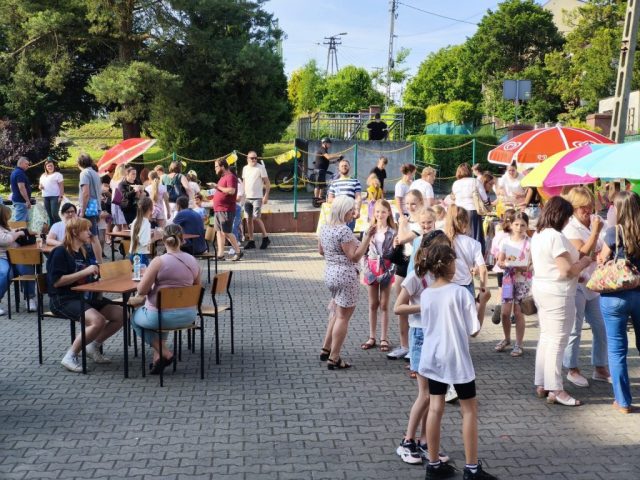 Uczniowie i społeczność lokalna na placu szkolnym podczas rodzinnego festynu. Na środku placu stoły z krzesełkami, zaś naokoło placu stoiska ze smakołykami.
