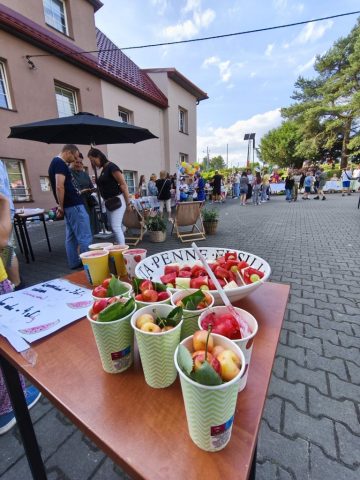 Uczniowie i społeczność lokalna na placu szkolnym podczas rodzinnego festynu. Widok na stoisko z owocowymi przekąskami - sałatka owocowa oraz świeże owoce w kubeczkach