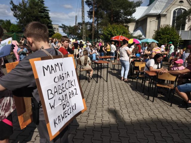 Uczniowie i społeczność lokalna na placu szkolnym podczas rodzinnego festynu. Na środku placu stoły z krzesełkami przy których siedzą uczniowie z rodzicami zajadając się owocowymi smakołykami. Na pierwszym planie uczeń z reklamą na plecach z hasłem: MAMY CIASTA I BABECZKI W SAM RAZ DO KAWECZKI.