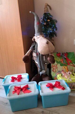 Na zdjęciu widoczne są trzy niebieskie plastikowe pojemniki ustawione na podłodze, każdy ozdobiony dużą czerwoną kokardą. Za pojemnikami stoi duża pluszowa dekoracja w kształcie renifera w czapce i szaliku. Obok znajduje się mała choinka z lampkami oraz zapakowane prezenty. Całość tworzy świąteczną dekorację w pomieszczeniu przedszkolnym.