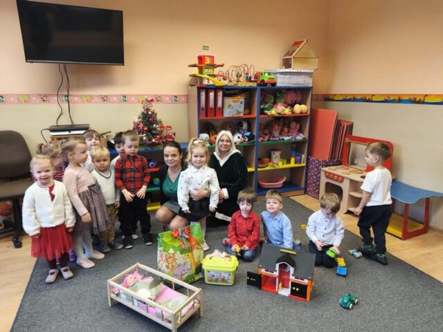 Na zdjęciu widać grupę dzieci oraz dwie osoby dorosłe, pani dyrektor ZSP 3 w Wodzisławiu Śląskim oraz pani sekretarz, pozujące razem w sali przedszkolnej. Dzieci stoją i siedzą wokół dorosłych. Przed nimi ustawione są zabawki, w tym domek dla lalek oraz inne akcesoria. W tle znajduje się mała udekorowana choinka oraz półki z zabawkami. Uczestnicy zdjęcia mają na sobie odświętne ubrania. Zdjęcie dokumentuje wspólne spotkanie świąteczne w przedszkolu.