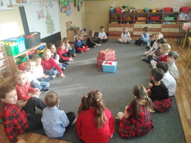 Na zdjęciu widać grupę około dwudziestu dzieci 5,6-letnich siedzących na dywanie w sali przedszkolnej. Dzieci siedzą w dużym kole, zwrócone twarzami do środka. W centrum koła znajdują się zapakowane prezenty w kolorowy papier oraz niebieskie plastikowe pojemniki z czerwonymi kokardami. Dzieci mają na sobie odświętne ubrania, część w kolorach czerwonym i białym. W tle widoczne są meble przedszkolne z zabawkami oraz świąteczne dekoracje na ścianach.
