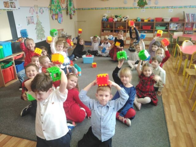 Na zdjęciu znajduje się grupa dzieci w sali przedszkolnej. Dzieci siedzą i klęczą na dywanie, trzymając w górze kolorowe klocki konstrukcyjne w kolorach zielonym, żółtym, czerwonym i niebieskim. Większość dzieci uśmiecha się i pokazuje swoje budowle. W tle widoczne są półki z zabawkami oraz elementy wyposażenia sali. Zdjęcie przedstawia moment wspólnej zabawy i prezentowania efektów pracy dzieci.