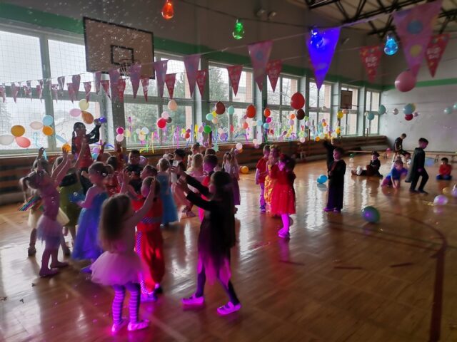 Dzieci tańczą w sali gimnastycznej w strojach bohaterów z bajek podczas baliku. Sala przystrojona jest kolorowymi balonami i błyszczącymi serpentynami.